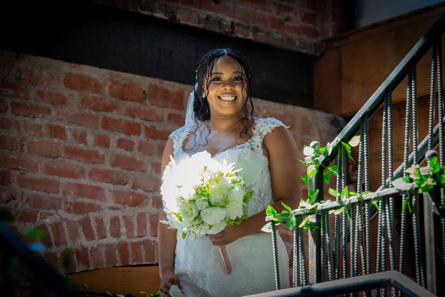 Smiling bride in white lace gown holding white bouquet on decorated staircase with brick wall backdrop.