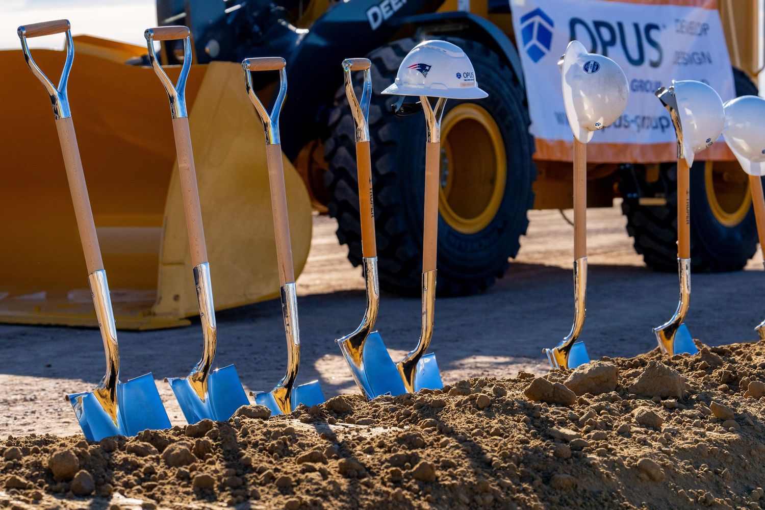 Row of ceremonial blue and gold shovels with white hard hats at an Opus Group groundbreaking event.