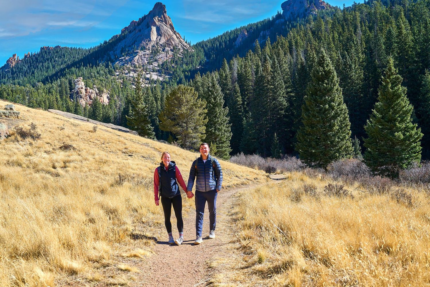 Two hikers walking together on a mountain trail with dramatic peaks in the background.