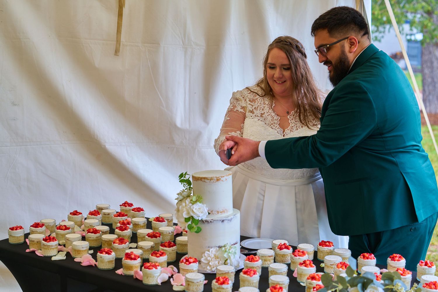 Newlyweds cutting their wedding cake together at dessert table filled with mini strawberry treats.