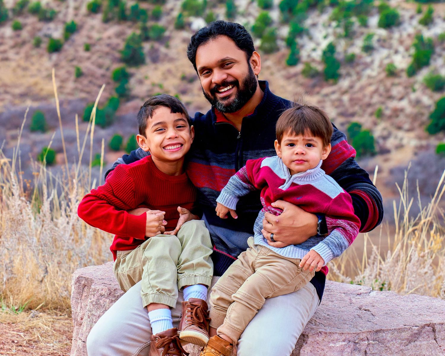 Father and two young children sit together on rocky terrain with mountain landscape behind them.