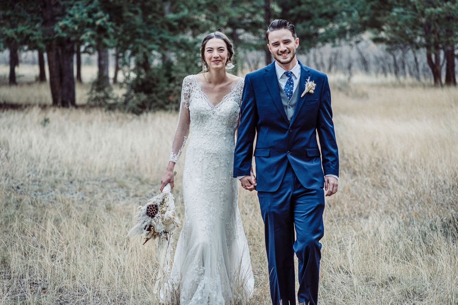 Newlyweds walking together in field with bride in lace gown and groom in navy suit.