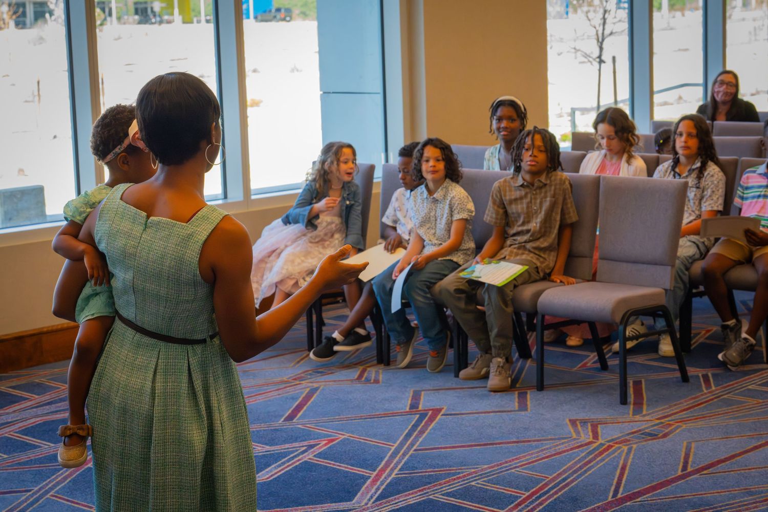 Woman in green dress presents to diverse group of students seated in chairs in a modern conference room.