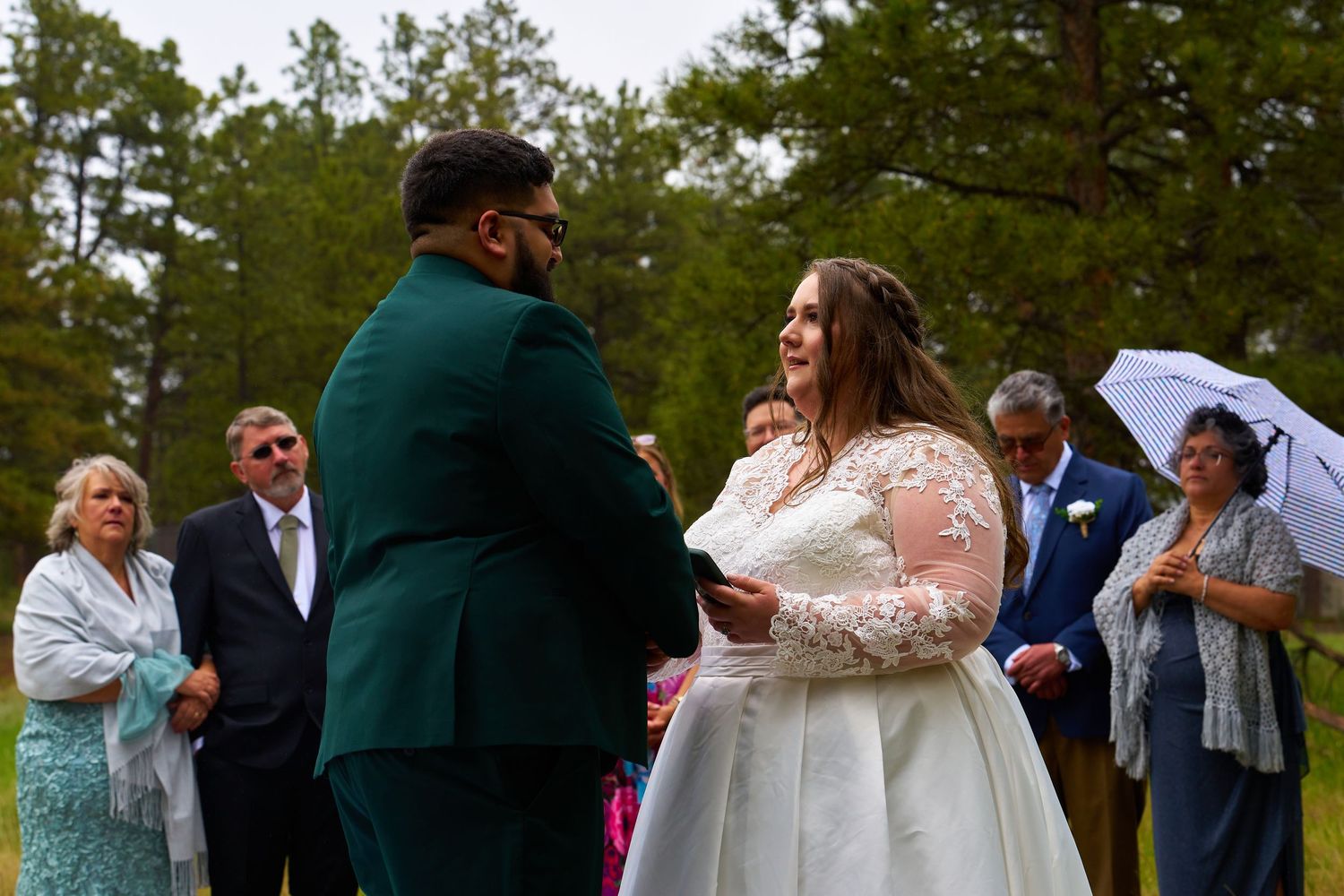 Outdoor wedding ceremony with couple exchanging vows surrounded by guests and pine trees.