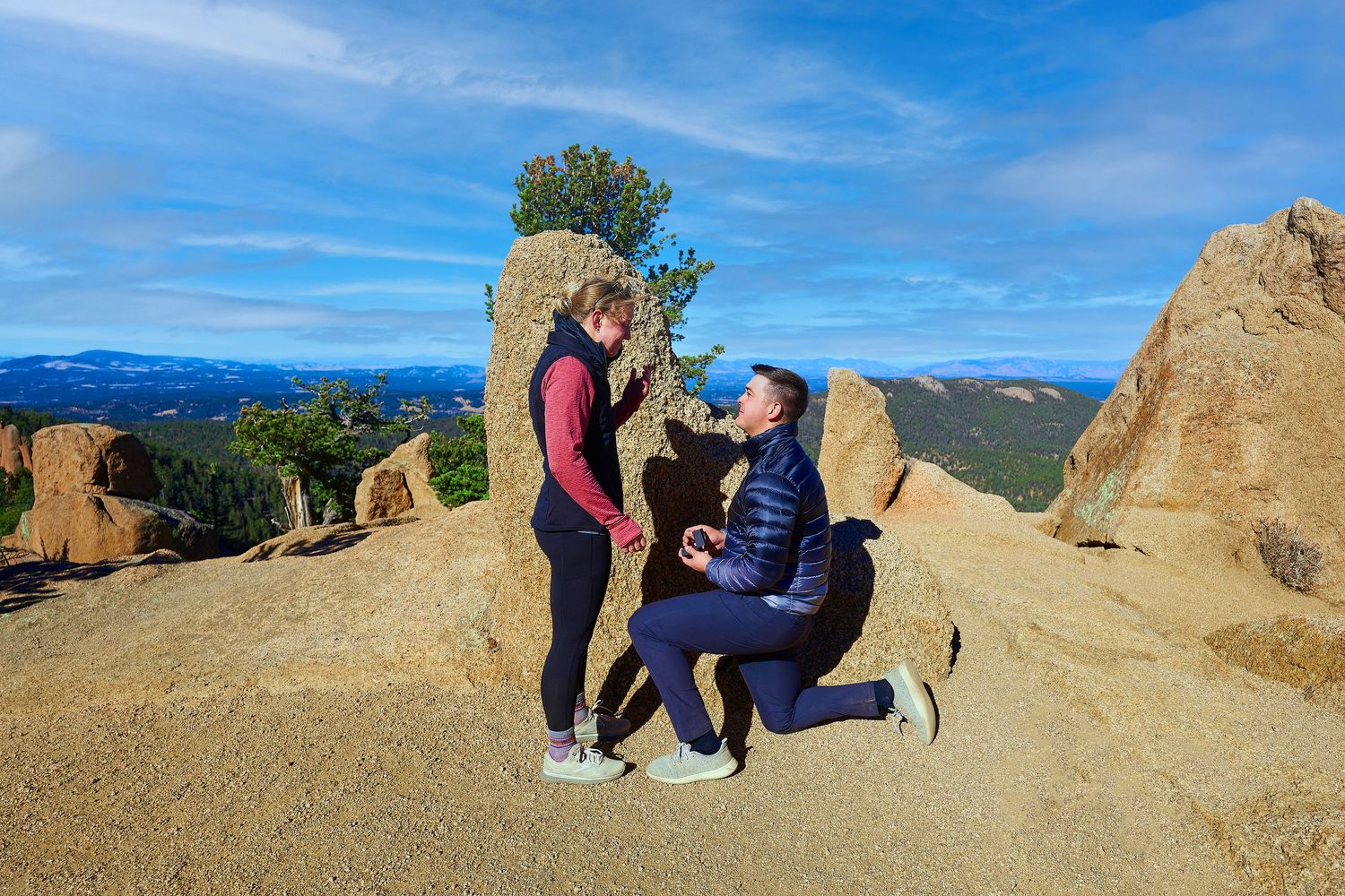 Couple sharing proposal moment on rocky mountain summit under blue sky.