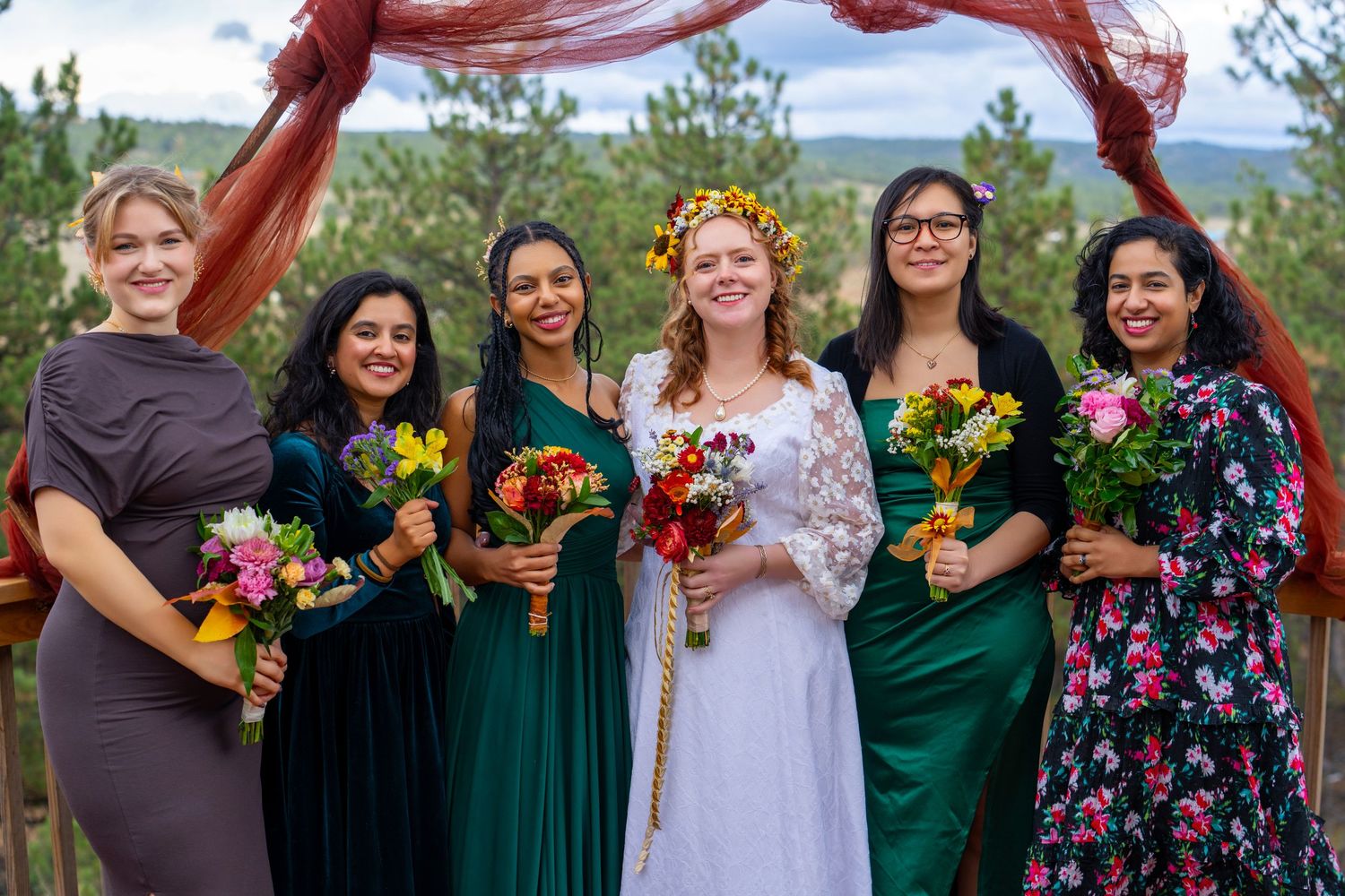 Wedding party in emerald green and floral dresses holds colorful autumn bouquets while red fabric flows in the wind.