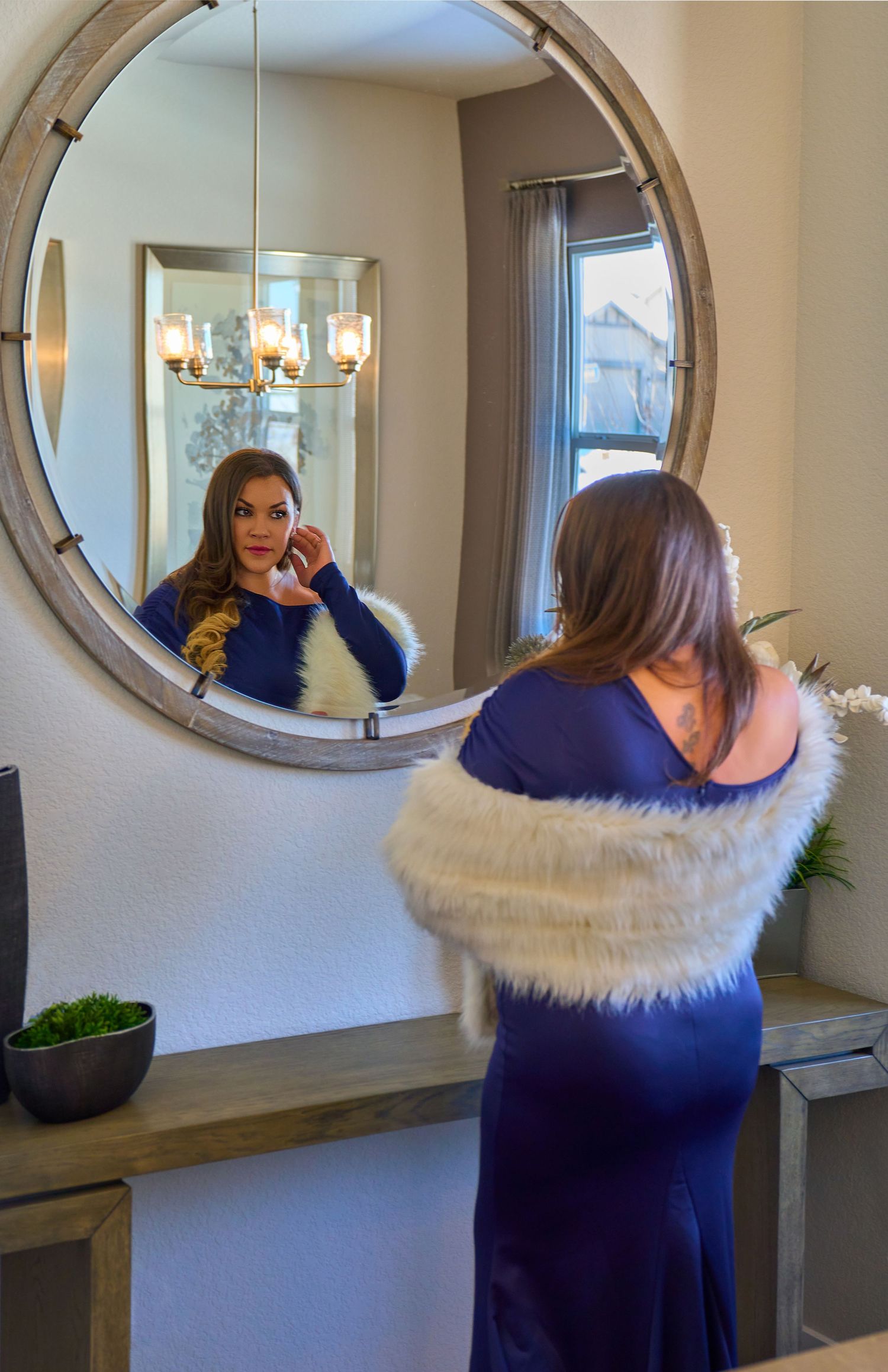 Person in a navy blue dress with white fur trim looks at their reflection in a round wall mirror near a chandelier.