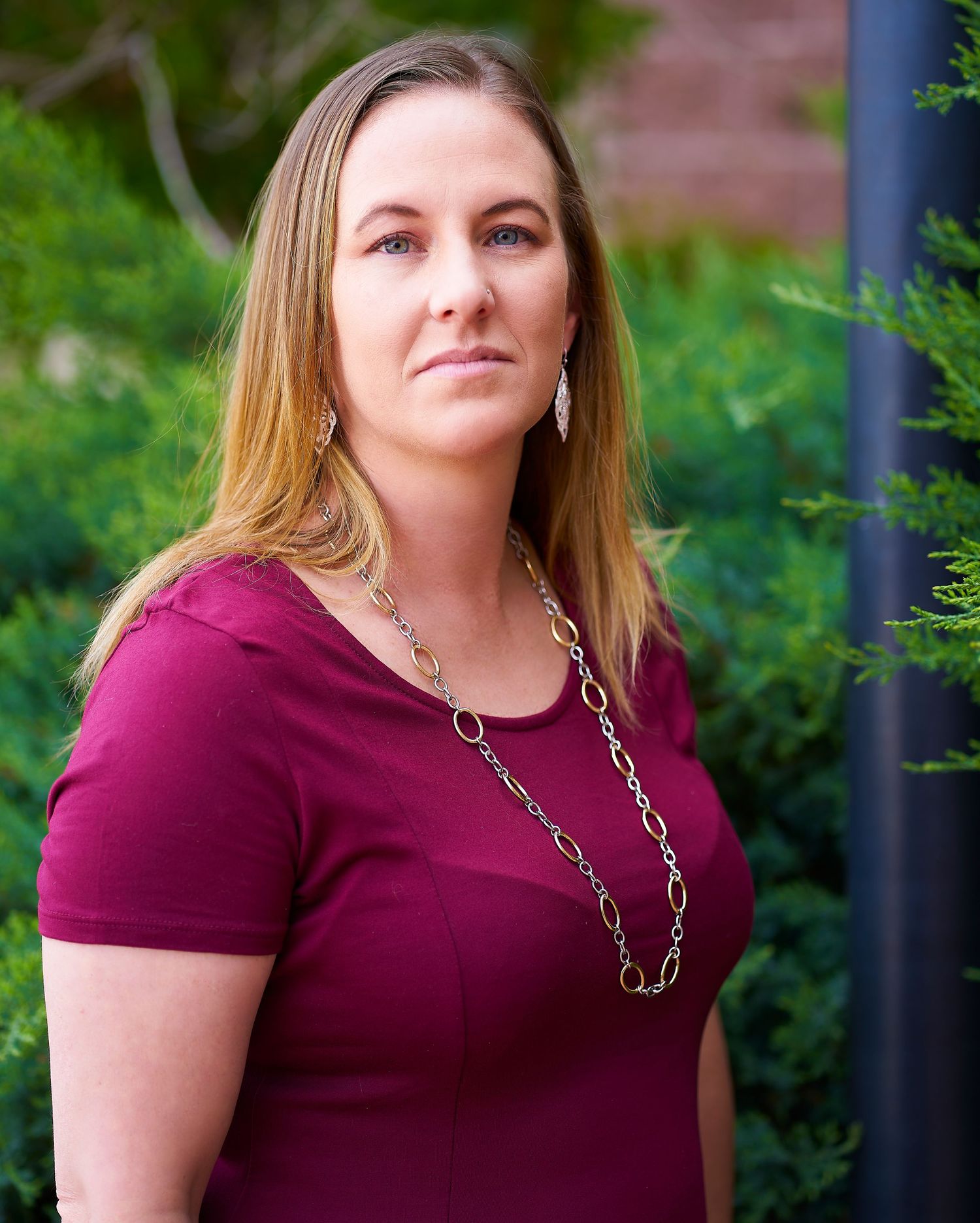 Professional in burgundy top with silver chain necklace poses outdoors against green foliage.