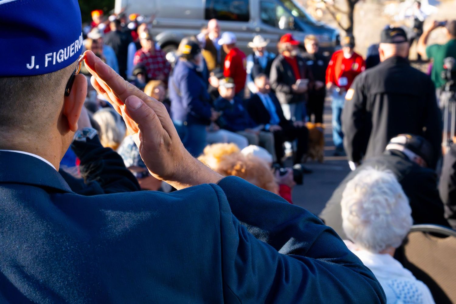 A uniformed figure salutes during an outdoor ceremony with a crowd gathered in the background.