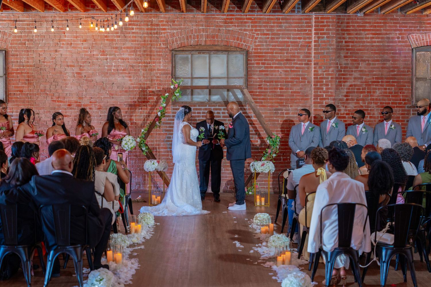 Elegant indoor wedding ceremony in a rustic brick venue with couple at altar, floral arch, candles, and seated guests.