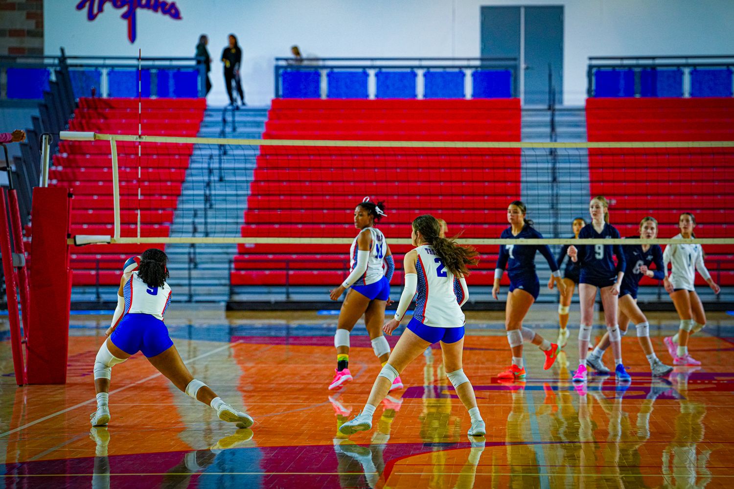 High school girls volleyball team practicing in a red and blue gymnasium with bleachers in the background.