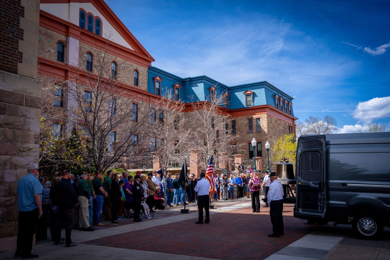 Crowd gathers outside historic red brick building during outdoor public event on a sunny day.