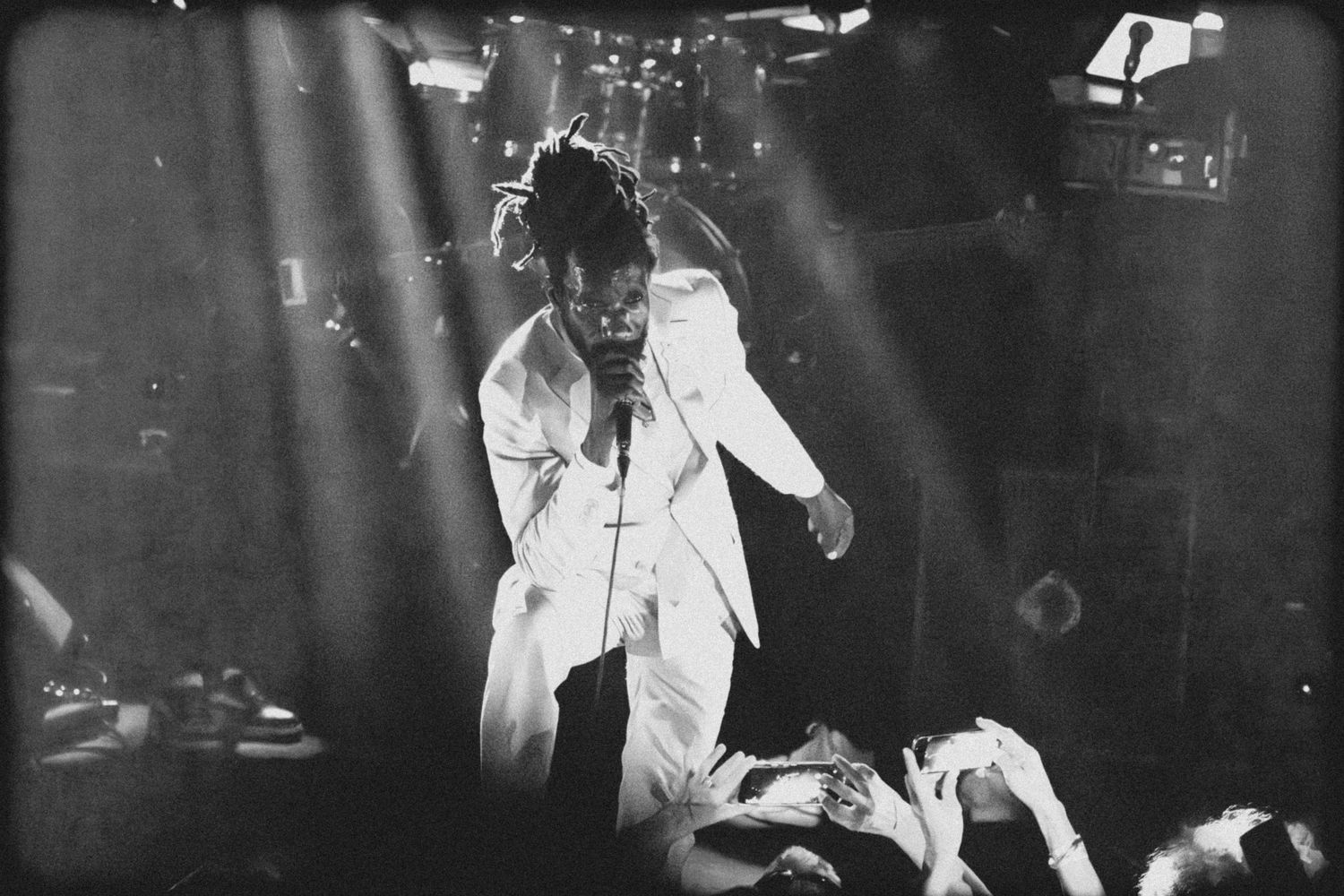Black and white photo of a performer in white clothing on stage during an energetic rock concert performance.