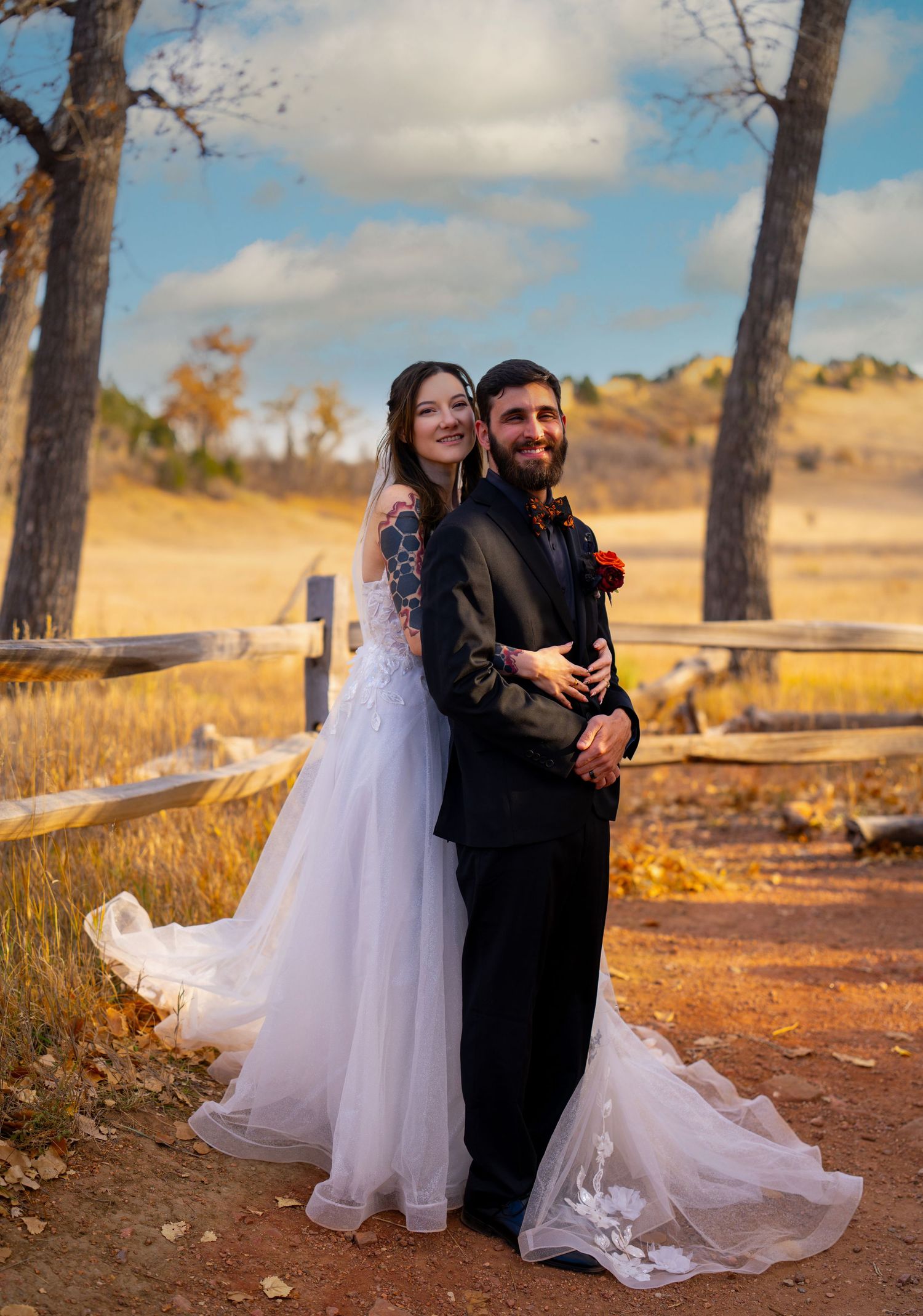 A couple in wedding attire poses romantically against a rustic wooden fence with autumn trees and hills in the background.