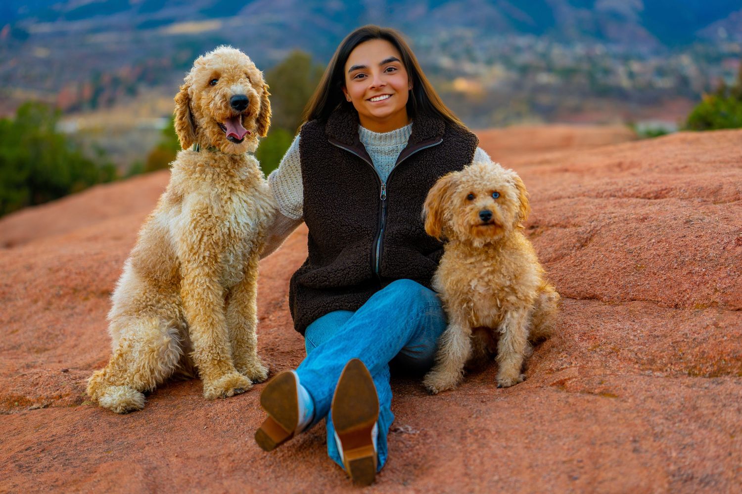 A person and their fluffy goldendoodle dog pose together on red rock formations in a scenic outdoor setting.