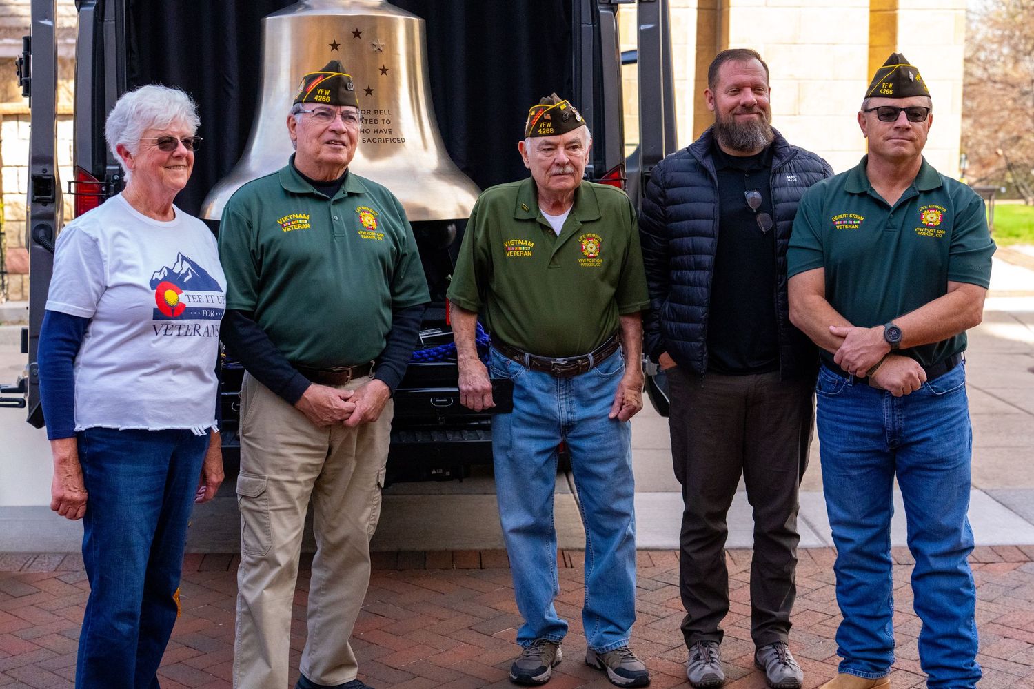 Five veterans pose together outdoors near a ceremonial bell during a military memorial event.