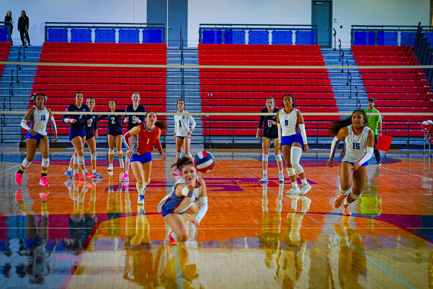 Female volleyball players compete on an indoor court with red bleachers, one player diving to dig the ball during a match.