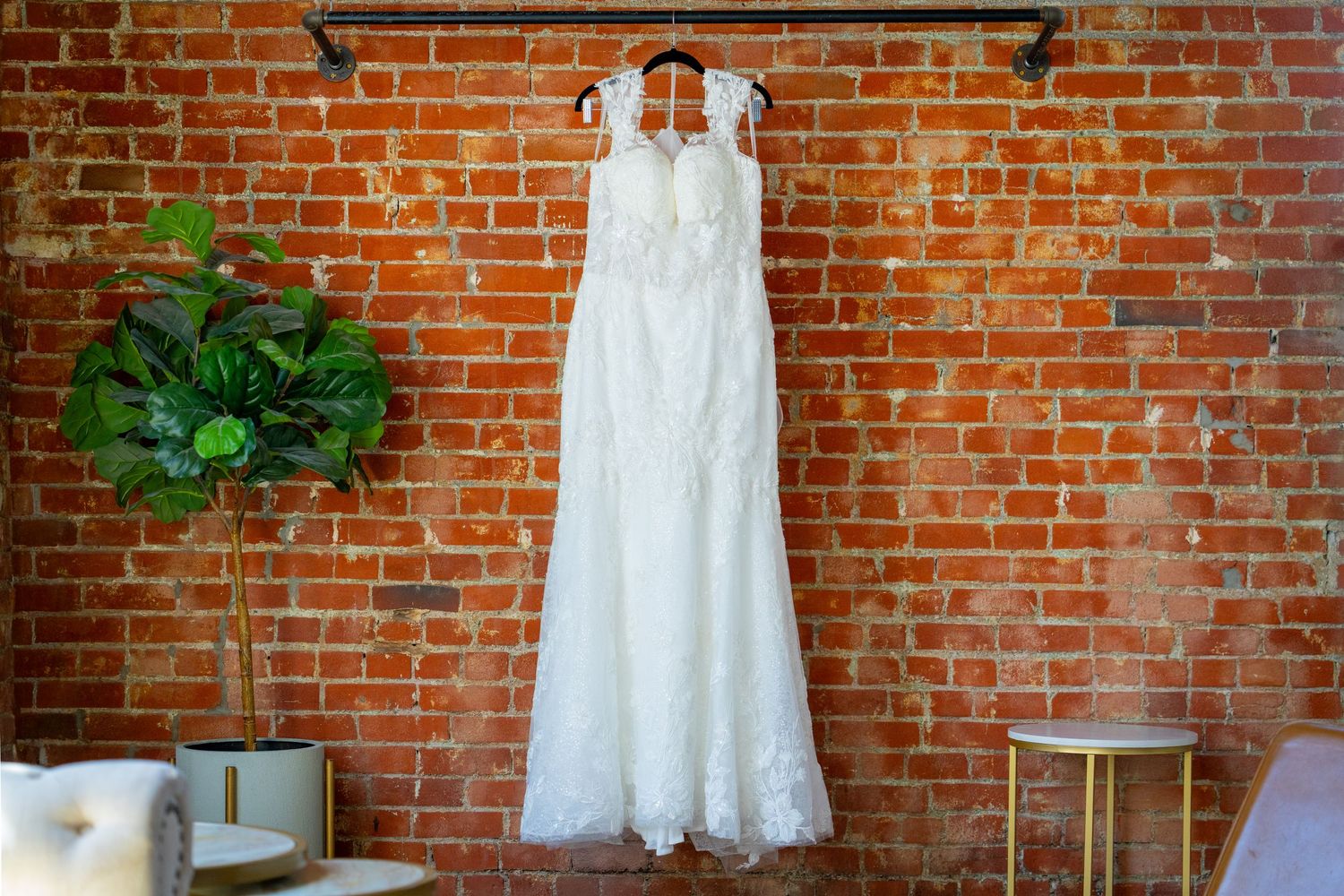 White lace wedding dress hanging on a rustic brick wall, displayed elegantly in a bright, stylish bridal boutique.