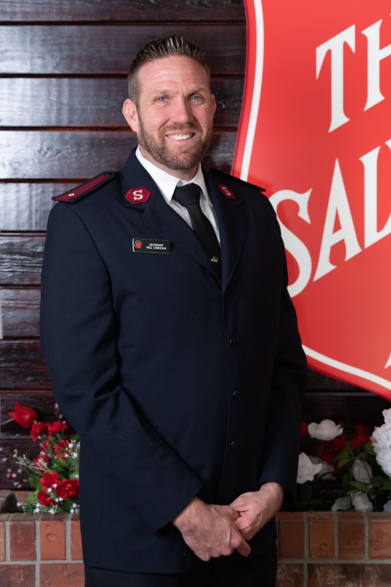 Salvation Army officer in formal uniform stands by organization logo and holiday decorations.