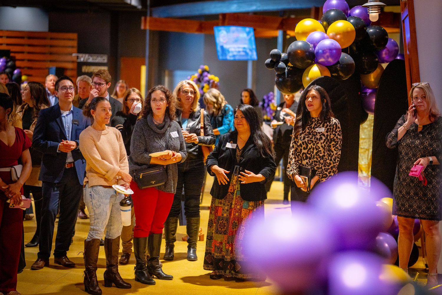 Guests gather at a festive event surrounded by purple, black, and gold balloons in a modern venue.