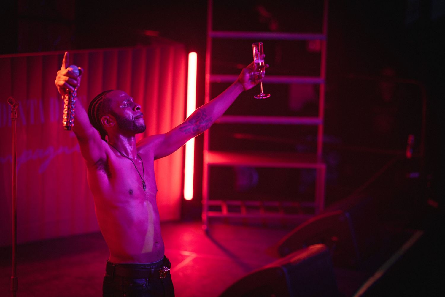 Shirtless man raising two drinks in a vibrant red-lit nightclub with neon lighting and a ladder backdrop.