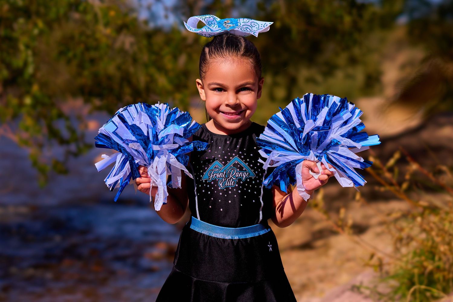 Young cheerleader with blue hair bow holds matching blue pom poms while performing outdoors.