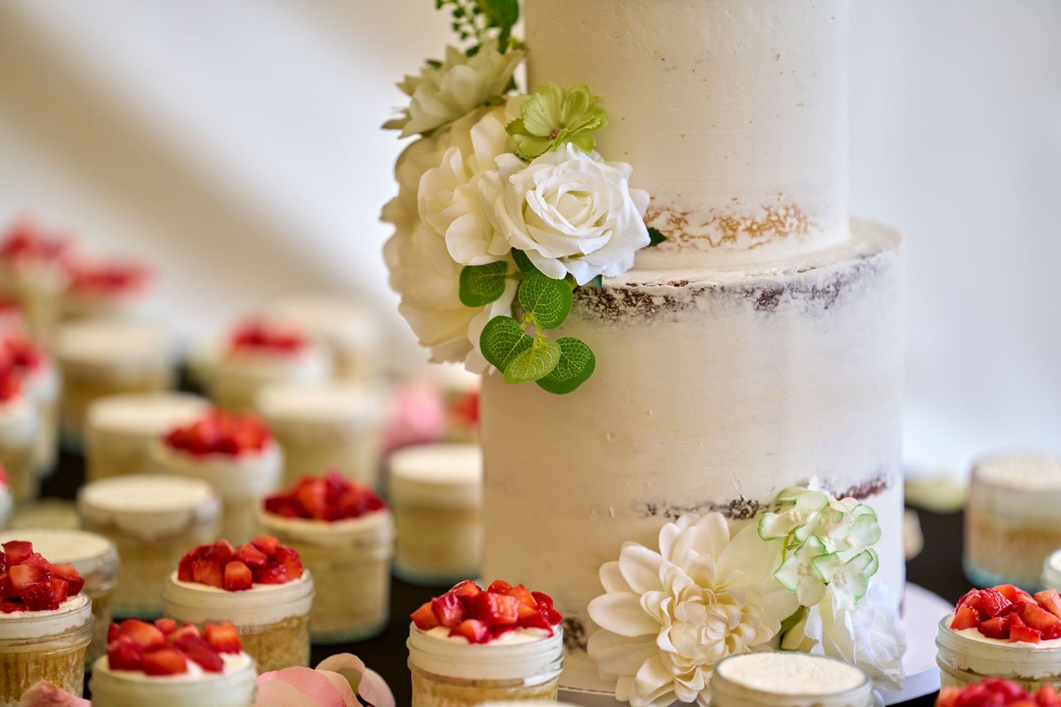 Elegant wedding dessert table with white tiered cake and mini strawberry topped desserts beautifully decorated with roses.
