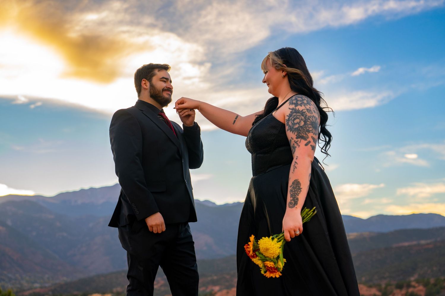 A couple shares a romantic moment at sunset against a backdrop of mountain peaks and dramatic clouds.