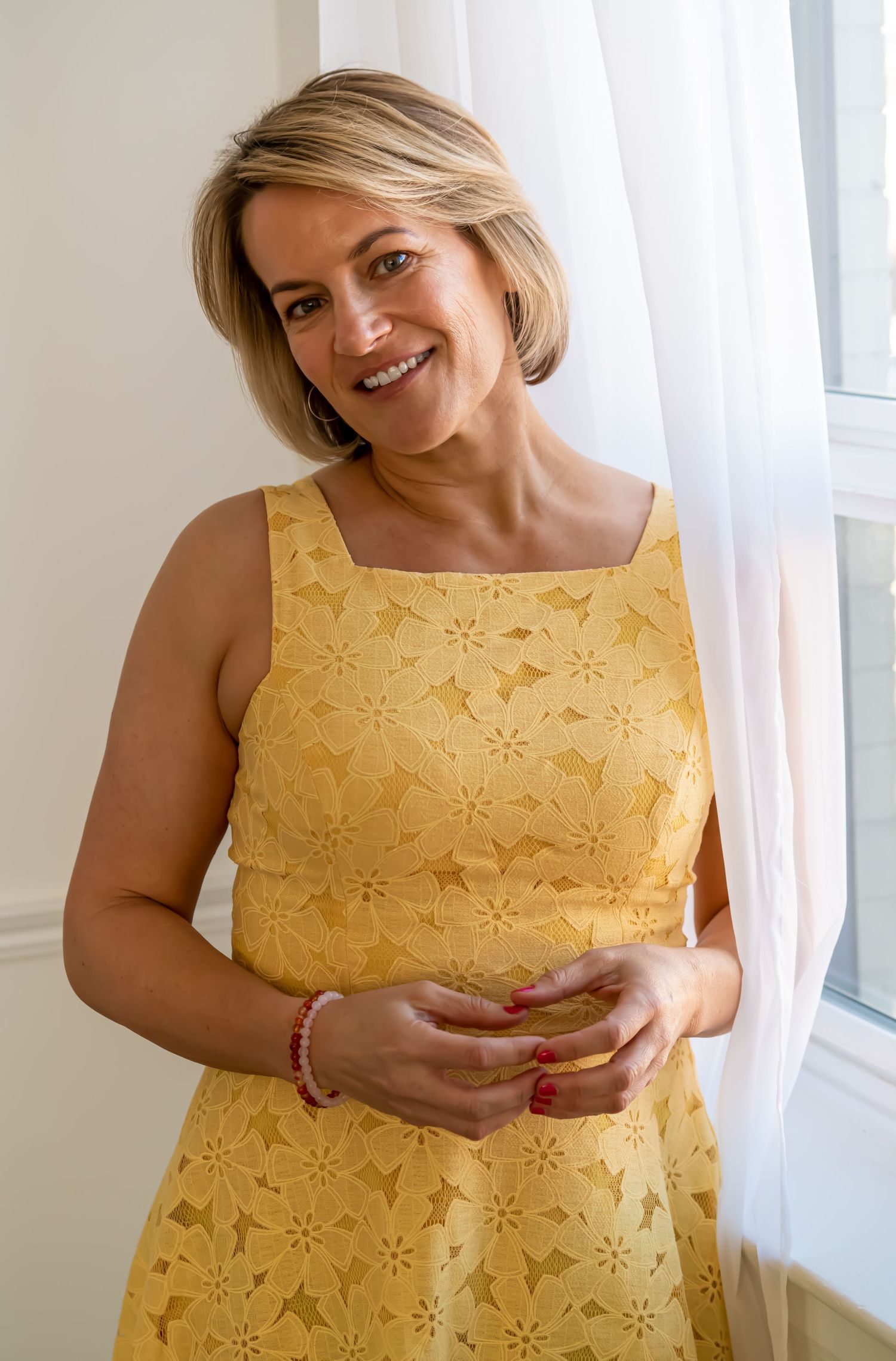 Woman in yellow floral lace dress smiling by window with natural light.