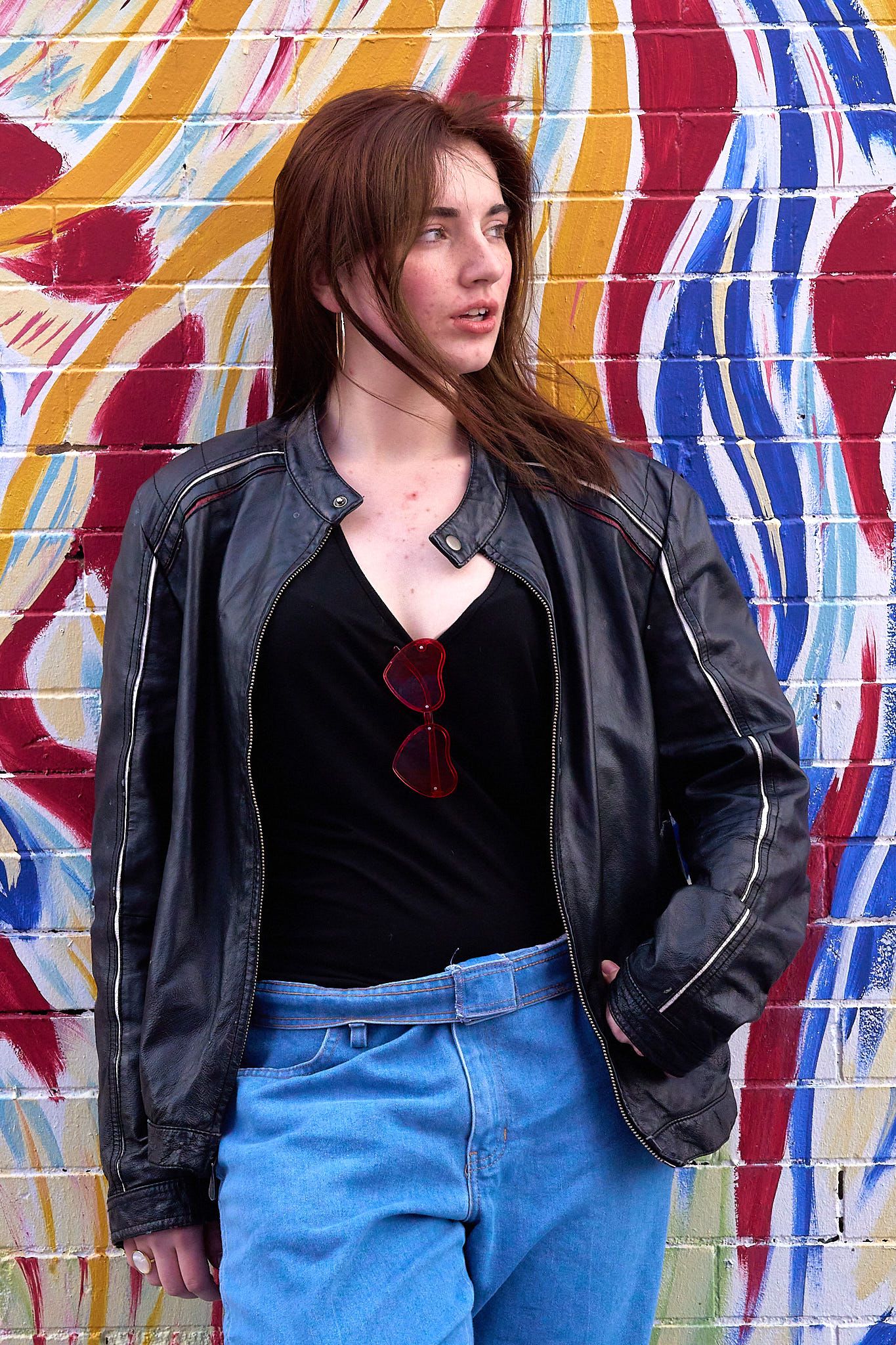 Person in leather jacket poses against colorful striped wall mural outdoors.