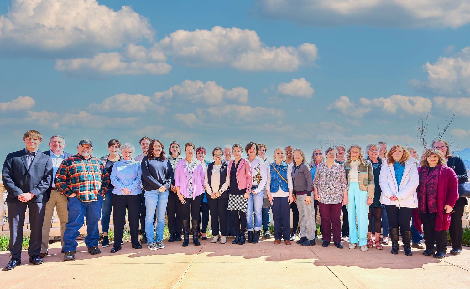 Large group photo of people standing together outdoors on a sunny day with blue sky and clouds.