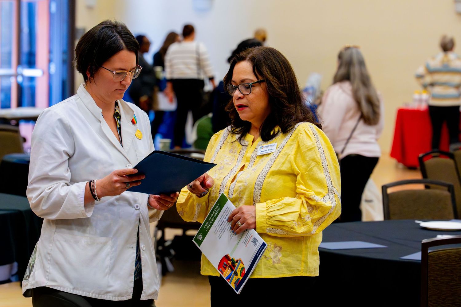 Two professional women reviewing documents together at a conference event in a bright meeting hall.