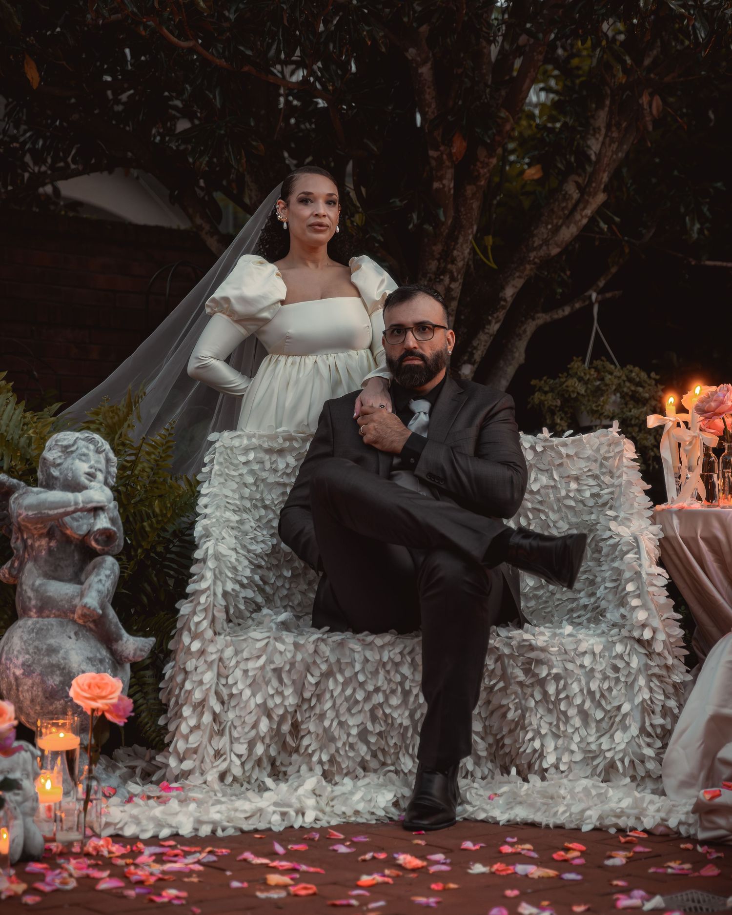 A couple in wedding attire playfully poses on a ruffled white couch surrounded by rose petals and candlelight.
