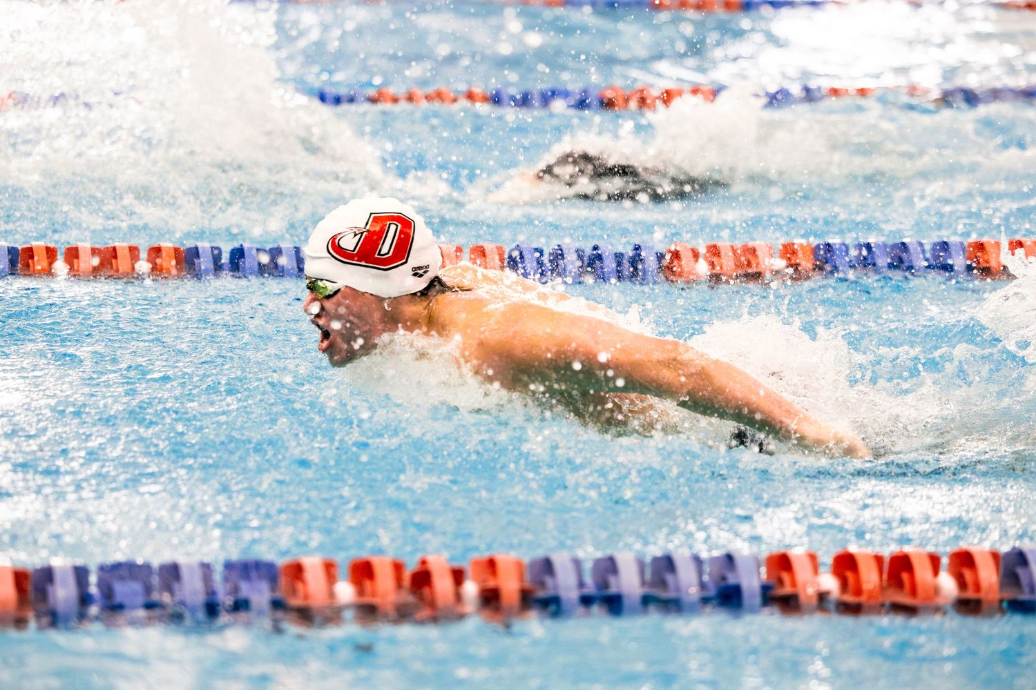 A swimmer performs butterfly stroke during a competitive race in a pool with red and blue lane dividers.