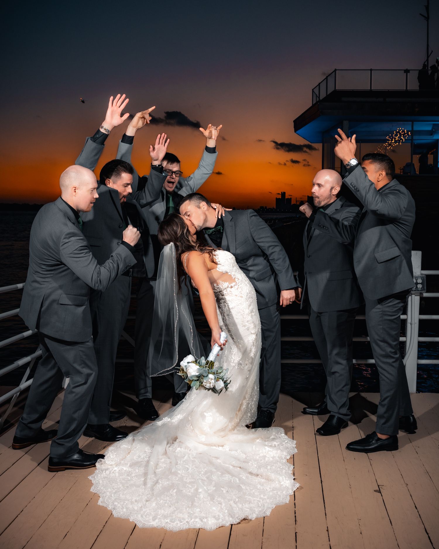 Wedding party poses playfully on a deck at sunset with dramatic orange sky in the background.