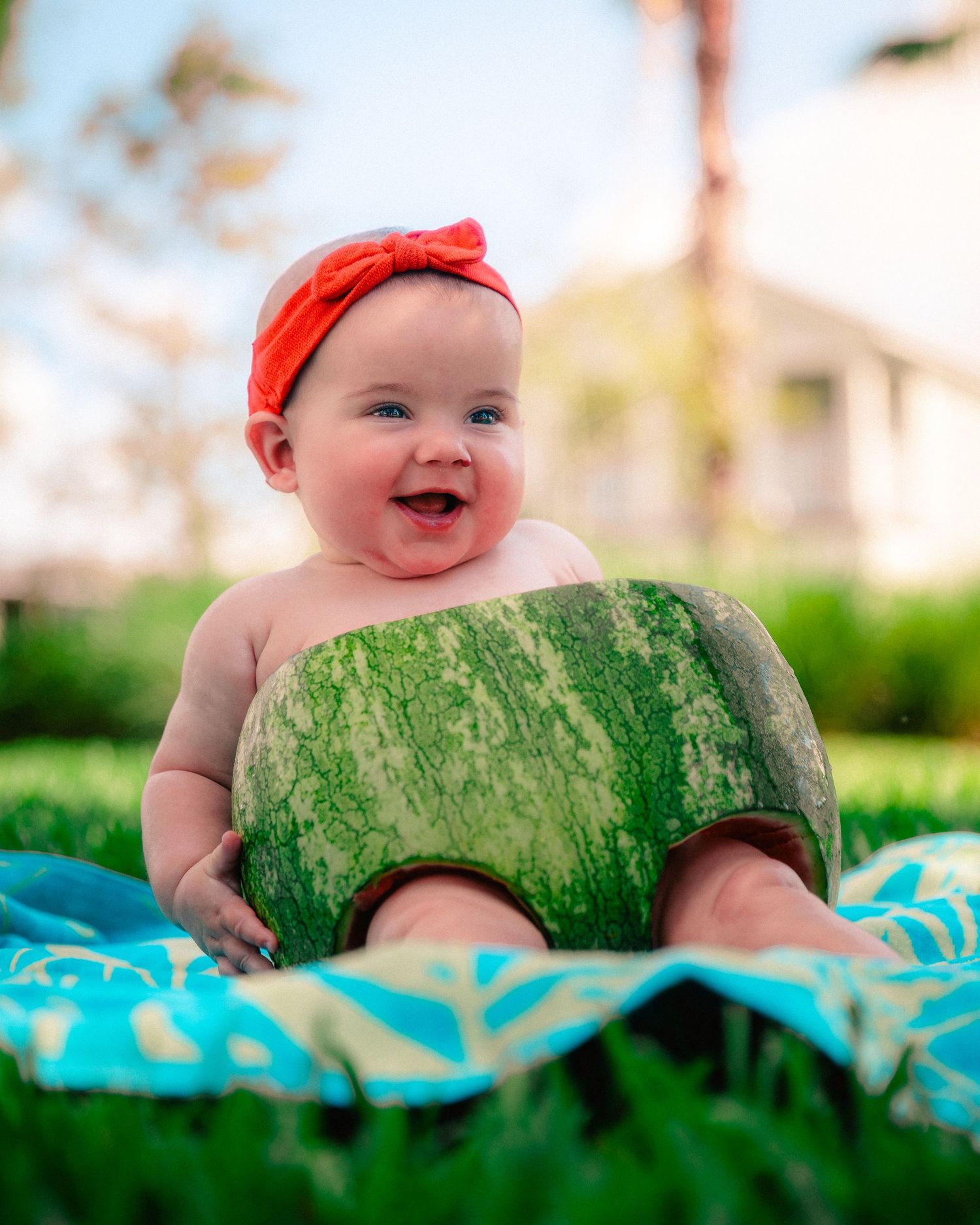 A baby wearing a red headband lies on a watermelon-themed blanket in a bright outdoor setting.