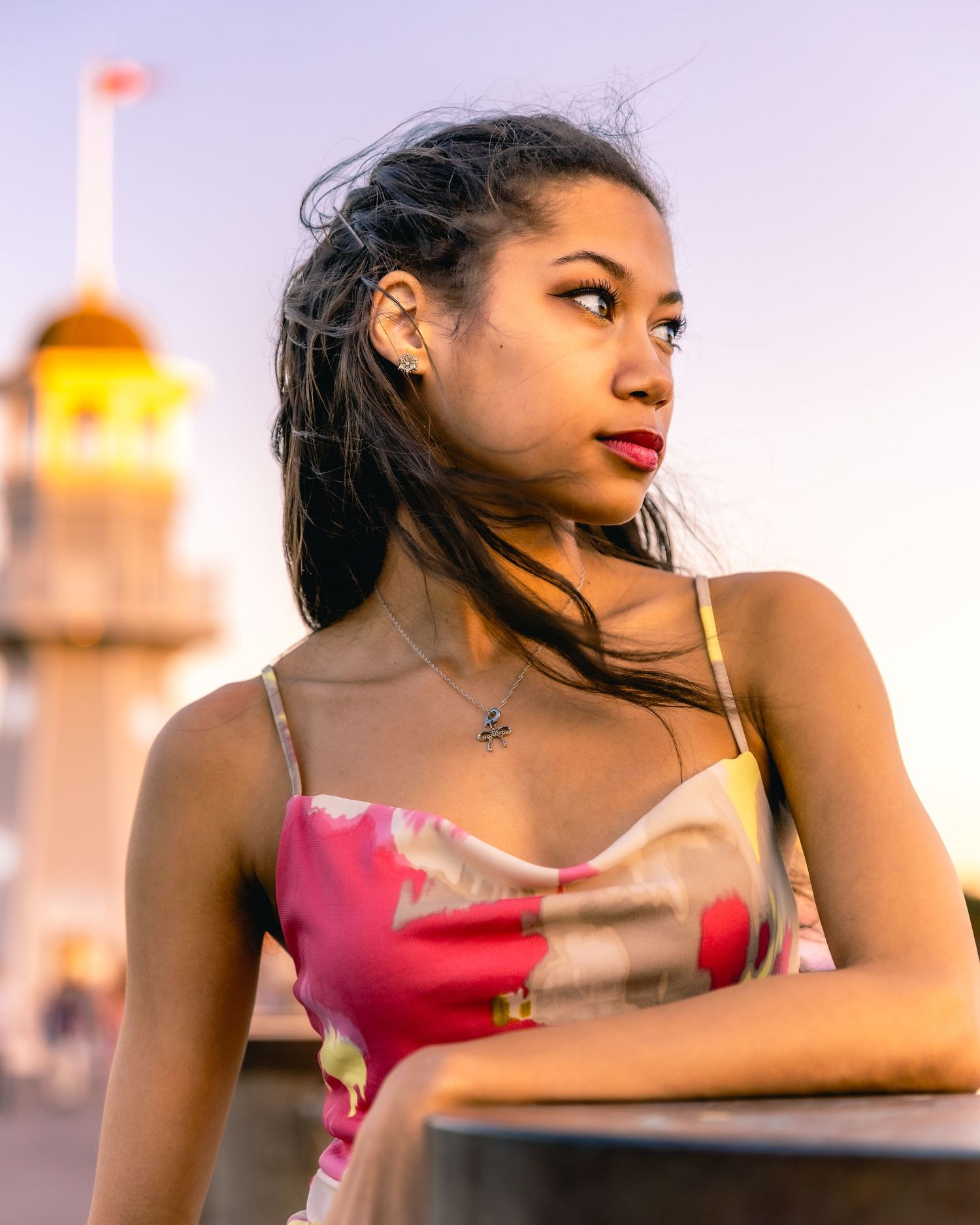 Young woman in pink dress poses thoughtfully against sunset backdrop with blurred lighthouse in distance.