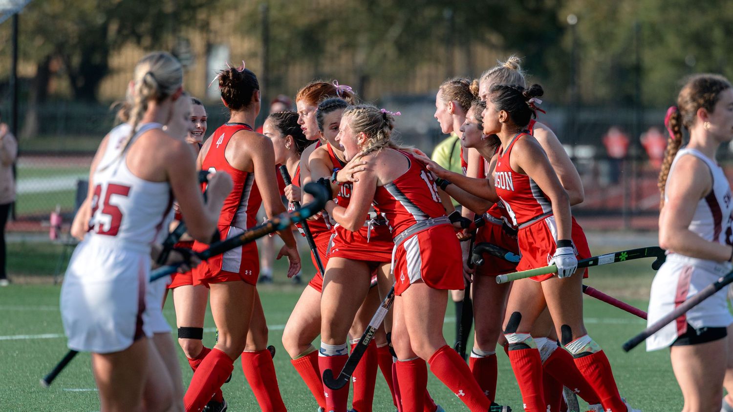 Field hockey players in red and white uniforms compete during an intense match on an outdoor turf field.