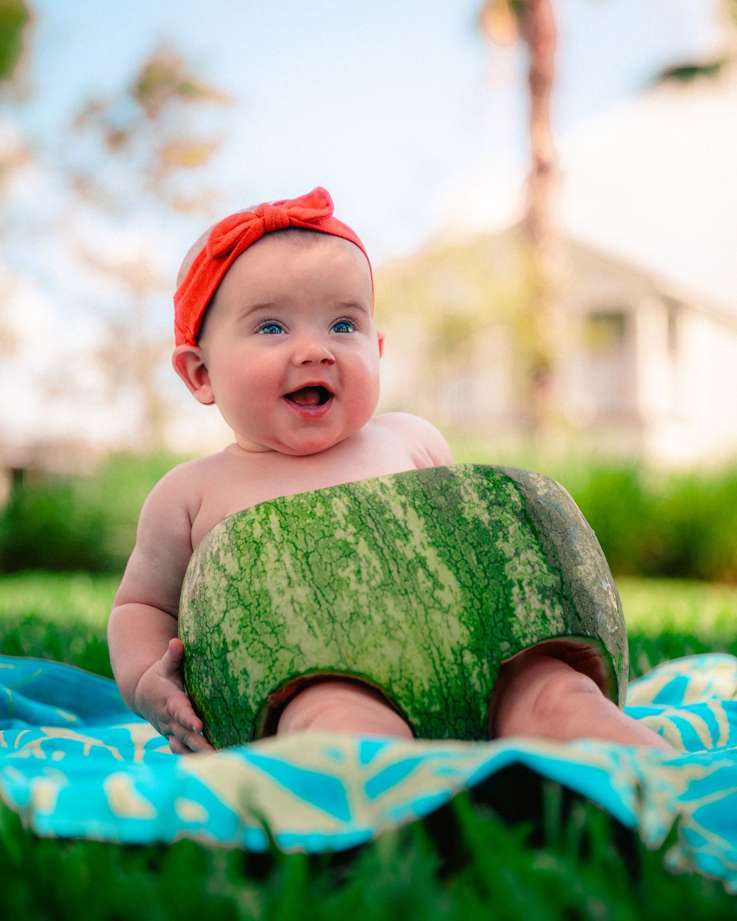 Baby wearing red hat lies on watermelon-patterned blanket in outdoor summer setting with blurred background.