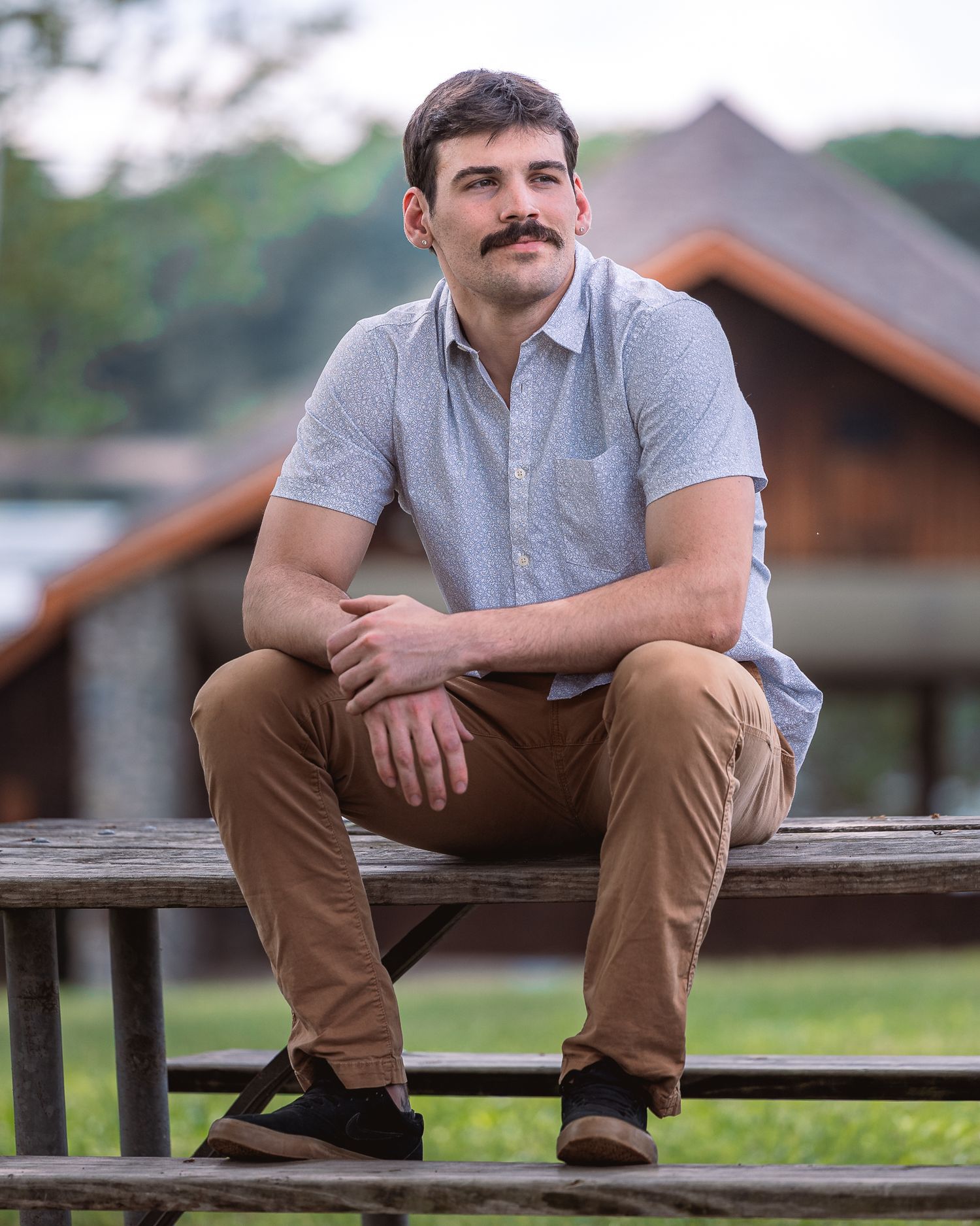 Person in casual attire sits thoughtfully on a wooden bench in an outdoor setting with blurred background.