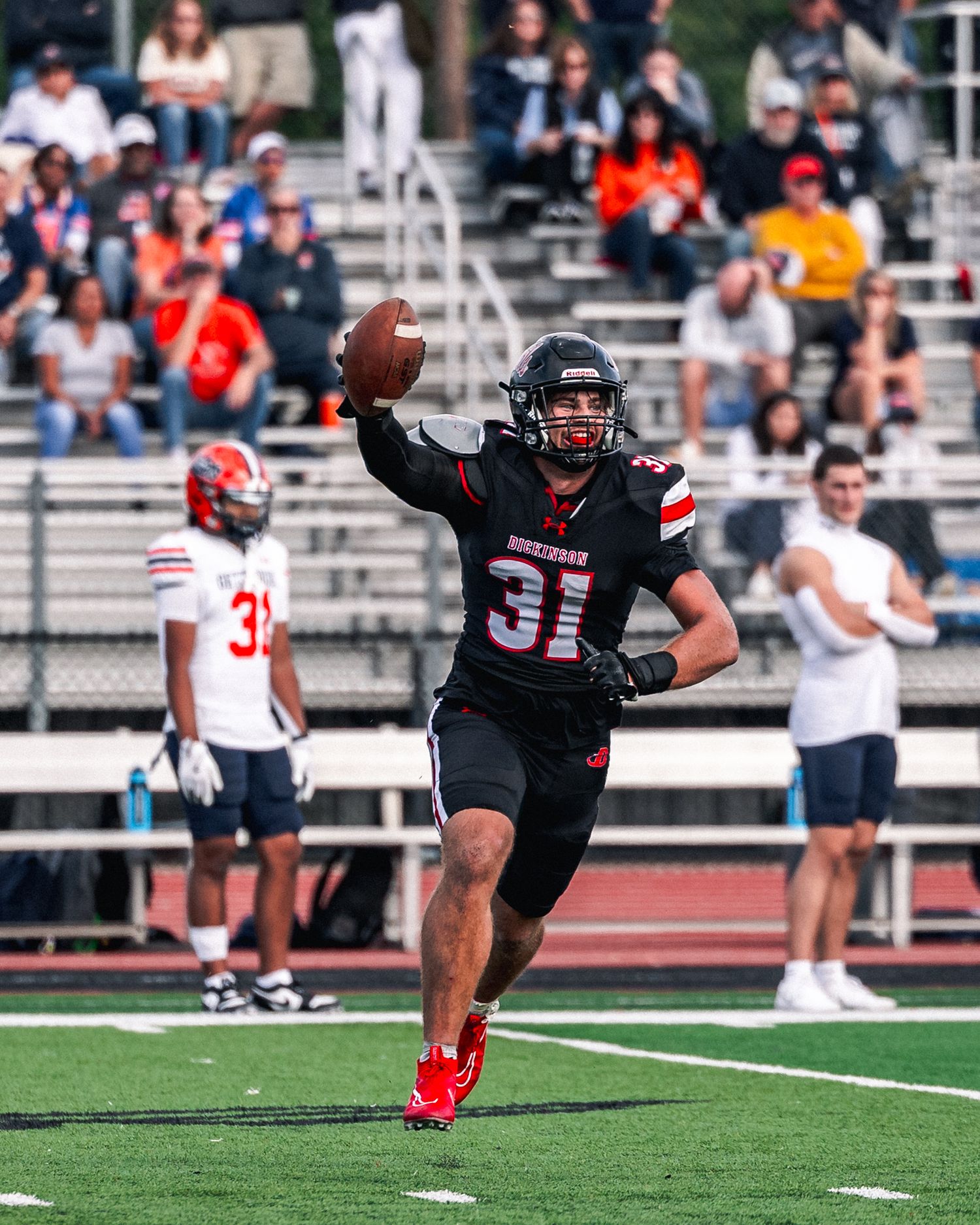 A football player in black uniform performs a kicking motion on the field during a game with spectators in the background.