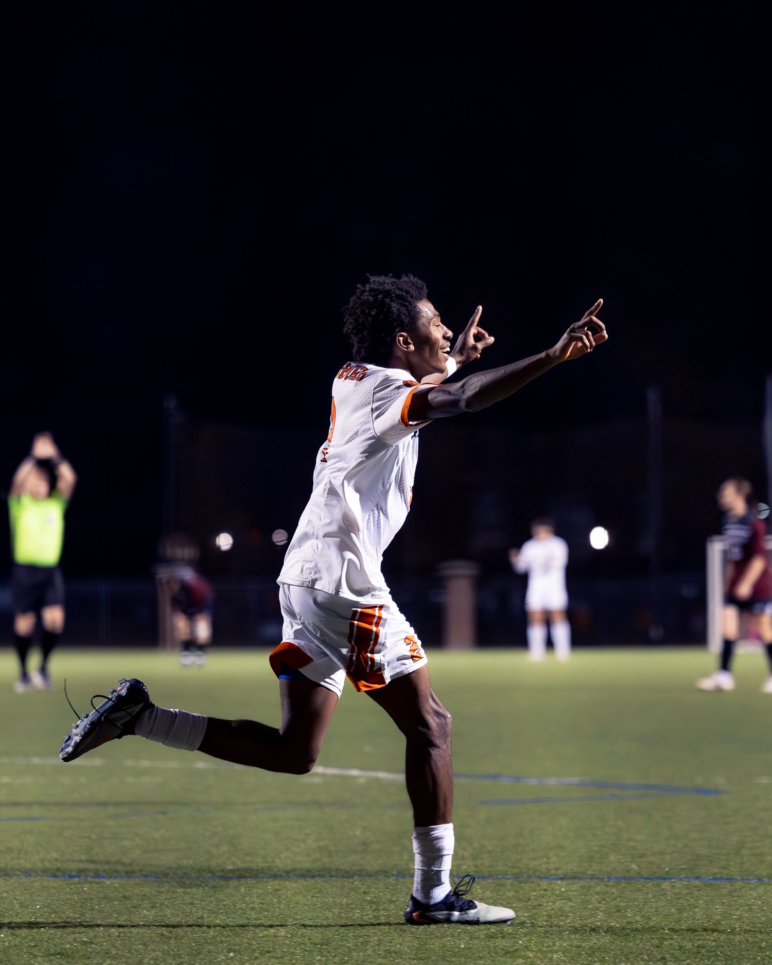 Soccer player celebrates by pointing and running with arms outstretched during nighttime match on turf field.