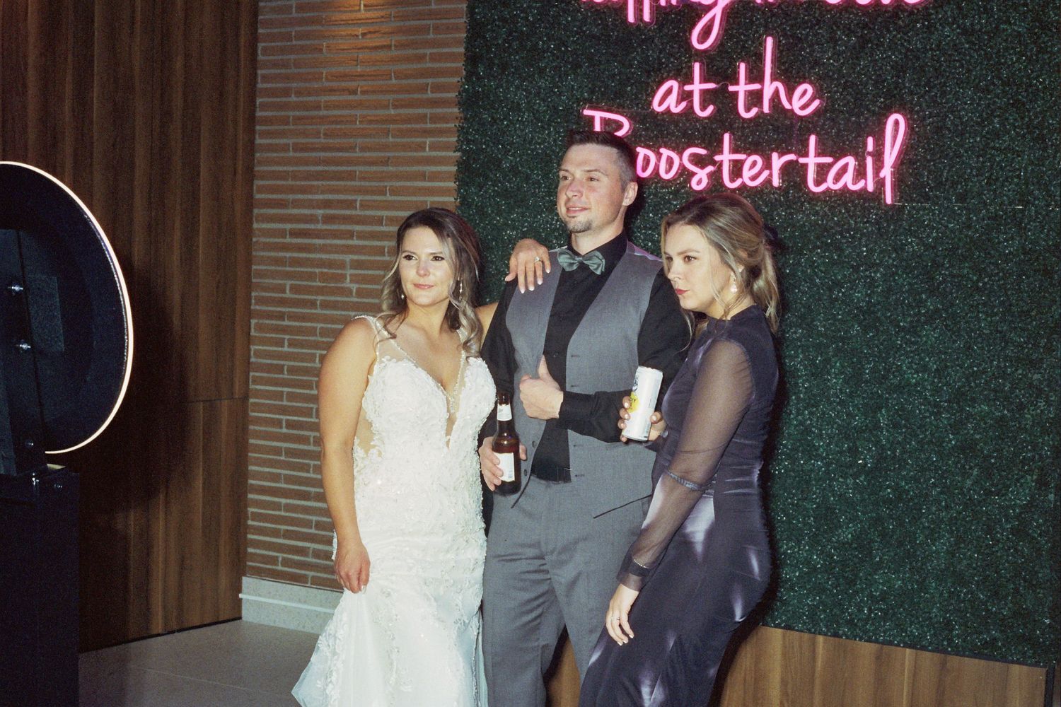 Three people pose together at an event with a pink neon sign and greenery wall backdrop.