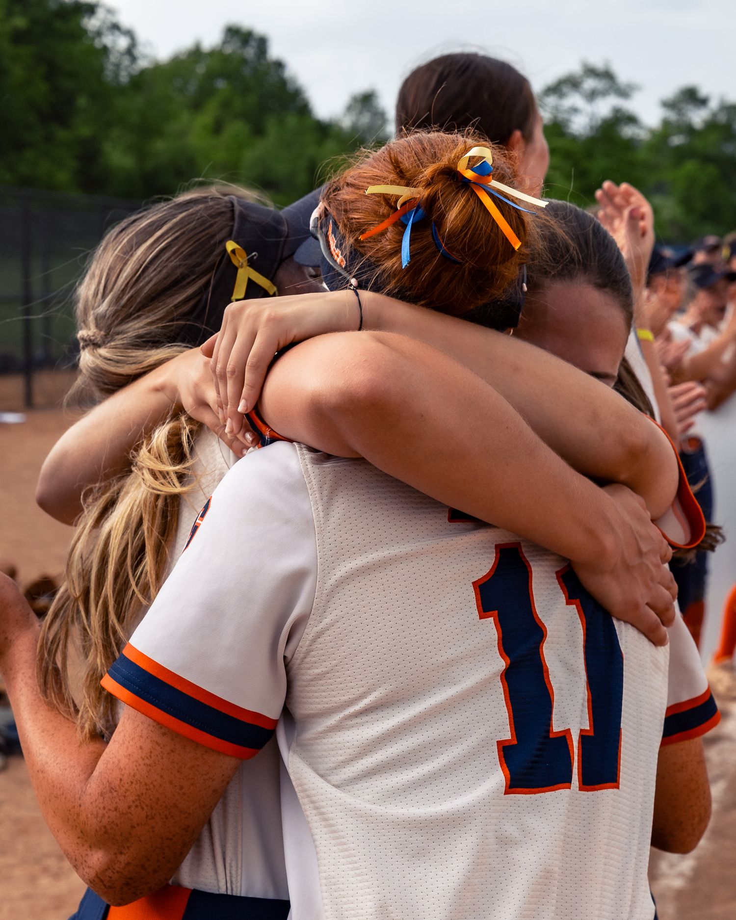 A group of athletes in white and blue uniforms huddle together in a supportive embrace on a sunny day.