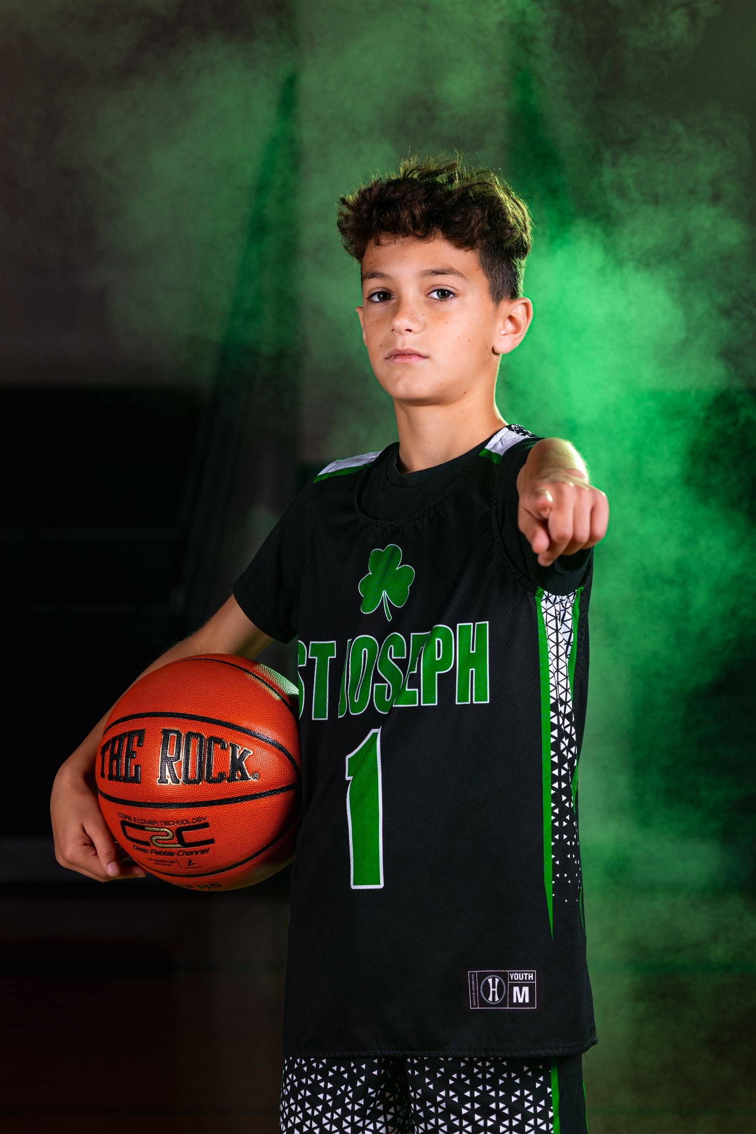 Youth basketball player poses in a green and black uniform with smoke effects in dramatic sports photography series.