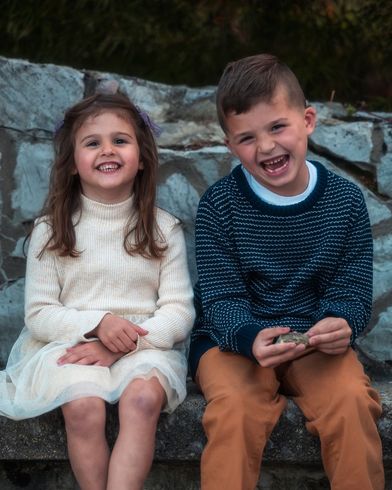Two children sitting together on stone steps share joyful laughter while wearing contrasting white and navy sweaters.