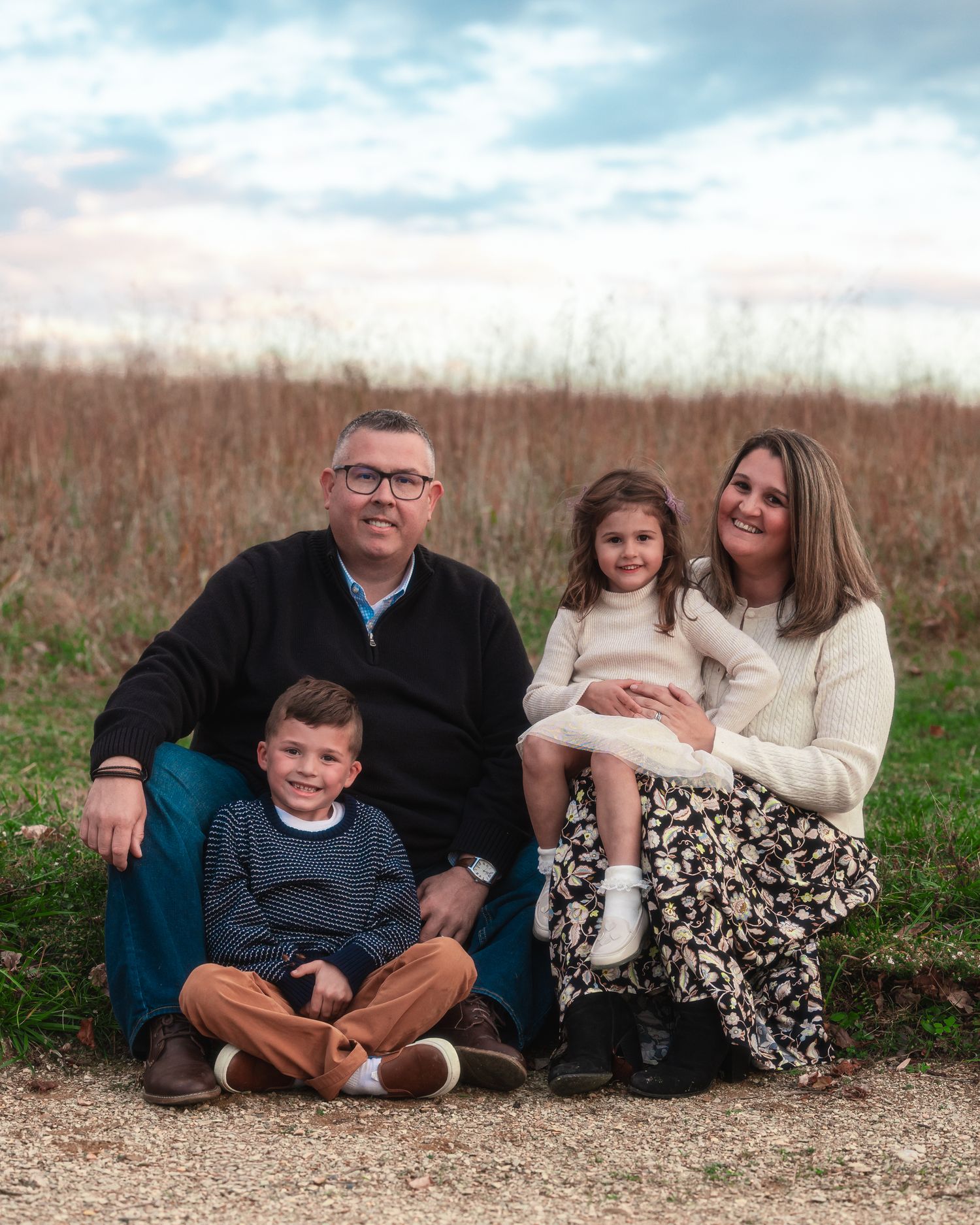 A family sits together outdoors on a gravel path with tall grass and cloudy skies in the background.
