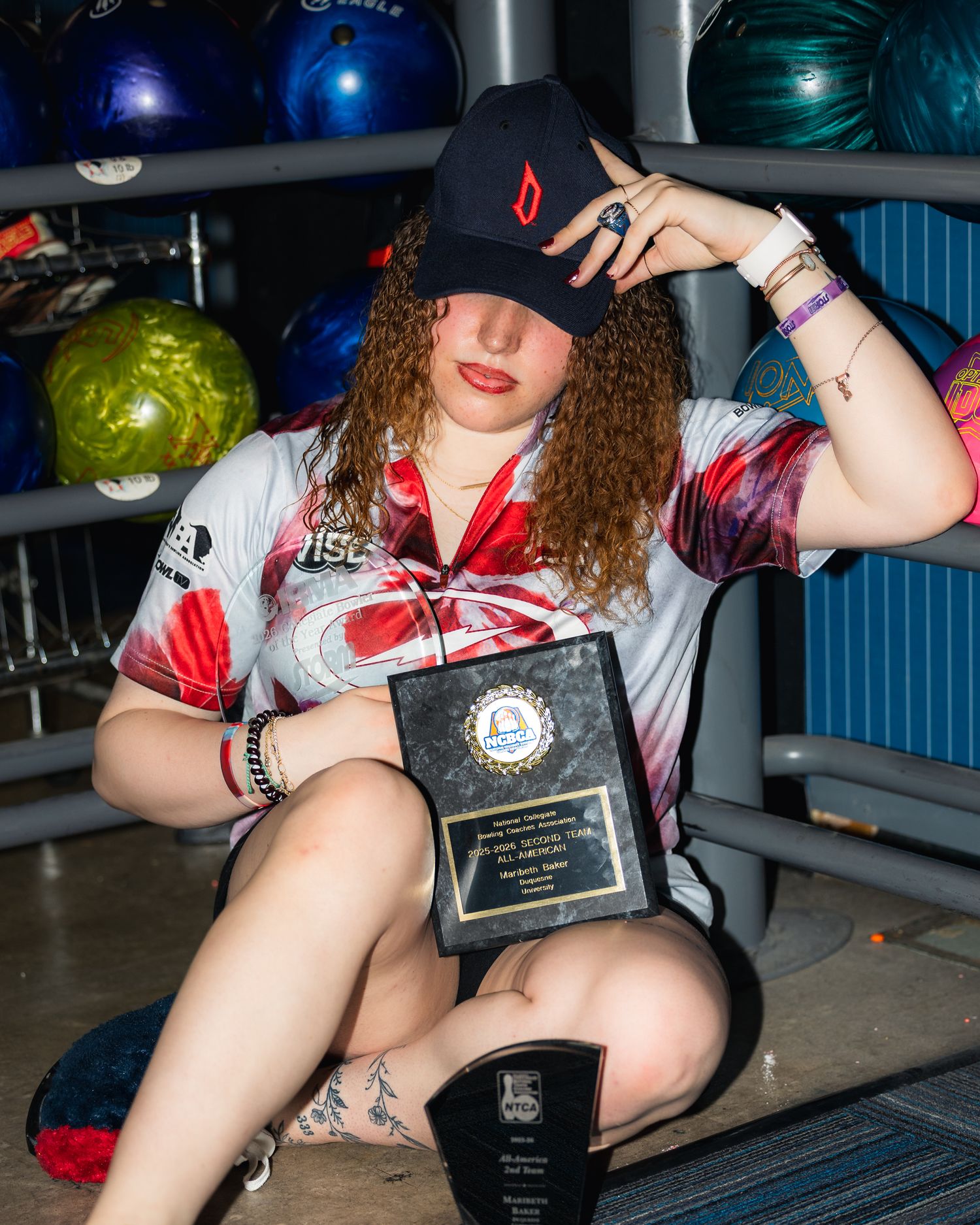 Young woman in bowling alley holding trophy plaque, wearing sport jersey and black cap, surrounded by colorful bowling balls.