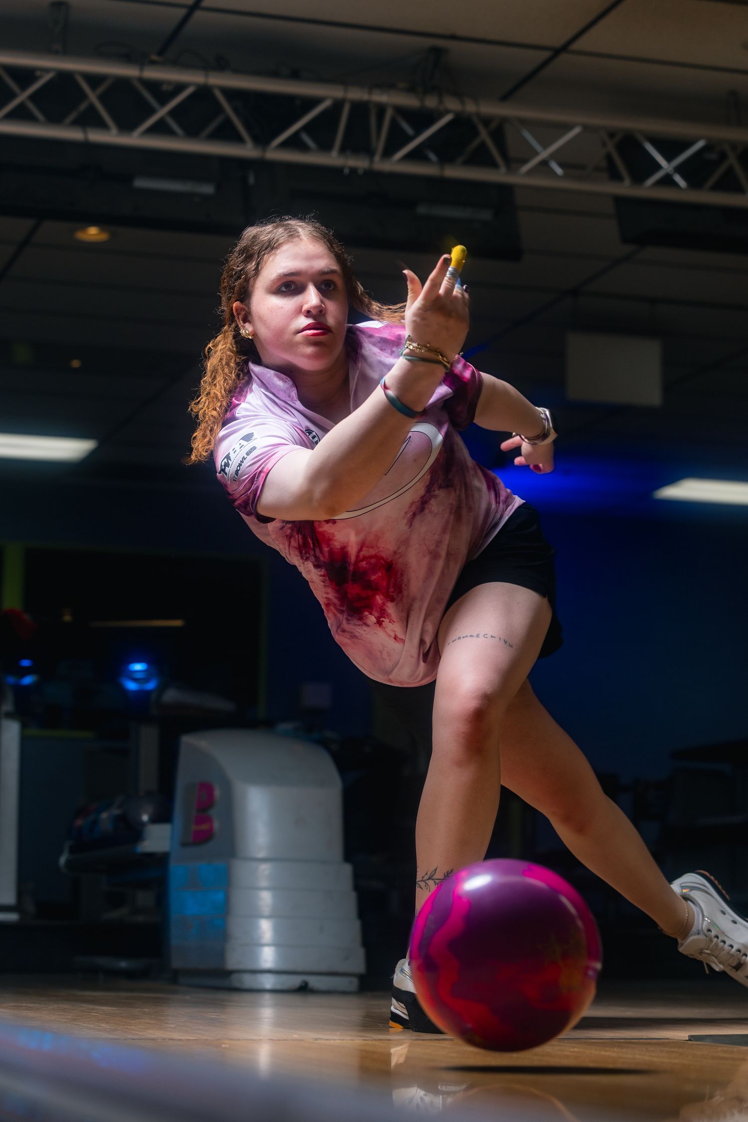 Young woman releasing a pink bowling ball on a lane in a bowling alley, wearing a tie-dye shirt.