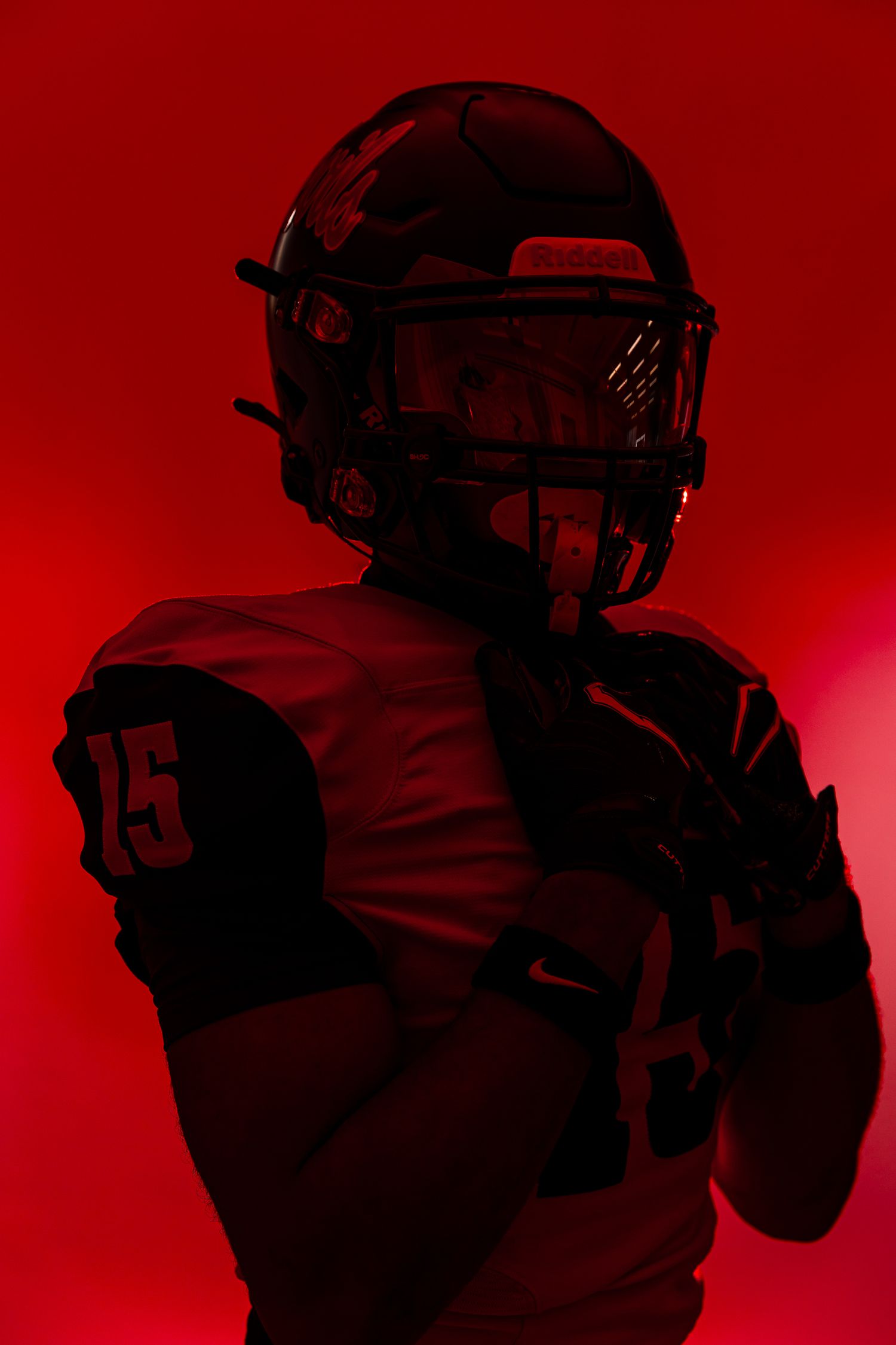 Football player in jersey number 15 poses dramatically against a bold red background wearing full helmet and gloves.